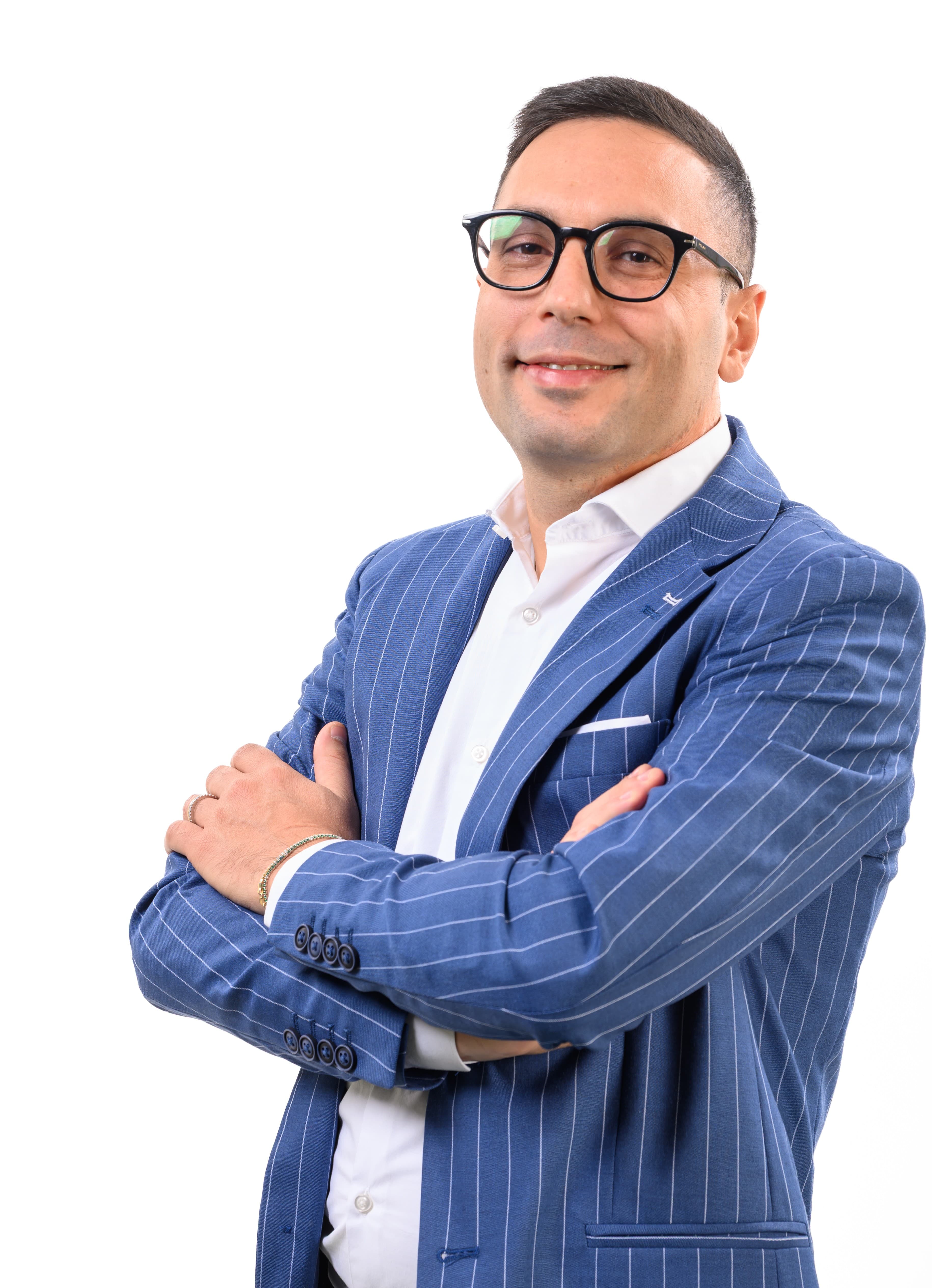 Uomo sorridente con occhiali, indossa una giacca blu gessata e camicia bianca, braccia incrociate.